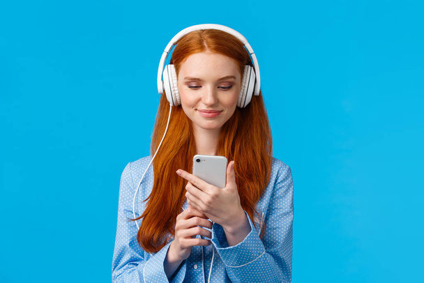 Περιμέντε-up πορτρέτο όμορφος foxy έφηβος κορίτσι κολέγιο με ακουστικά, ακούγοντας μουσική, κύλιση ειδήσεων το πρωί, τον έλεγχο των μέσων κοινωνικής δικτύωσης στο smartphone, στέκεται μπλε φόντο στην πιτζάμα - Φωτογραφία, εικόνα
