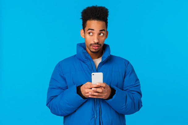 Thoughtful and creative cute african american guy thinking, use imagination to write cute message girlfriend, inviting come date valentins day, holding smartphone pensive, blue background - Fotografie, Obrázek