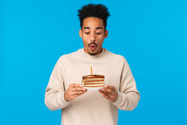Dovolená, koncepce oslav. Veselý Afroameričan b-den chlap slaví narozeniny, drží dort a sfouknout svíčky přání, stojící modré pozadí radostné, párty - Fotografie, Obrázek