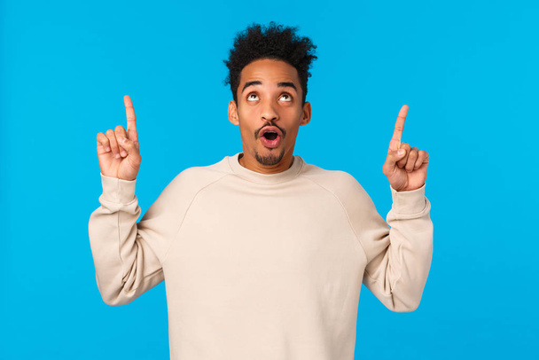 Wow awesome. Impressed good-looking african guy with moustache and afro haircut, folding lips smiling and staring, pointing up at amazing breathtaking event, advertising, blue background - Foto, Bild