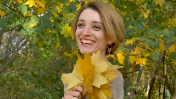 Emocional joven atractiva con cabello corto rubio y vestido biege sosteniendo un ramo de hojas de otoño por encima de su cabeza y posando en un hermoso parque al aire libre
 - Metraje, vídeo