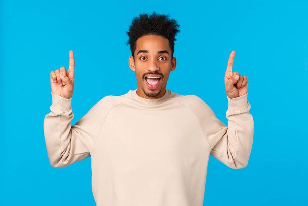 You gotta try this. Outgoing amazed and excited happy smiling african-american man loves winter holidays, attend awesome party, pointing fingers up, having fun and telling about promo, blue wall - Foto, Bild
