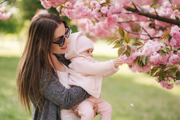 Mother show firs time to her daughter pink tree. Happy baby hold branch of sakura - Photo, Image