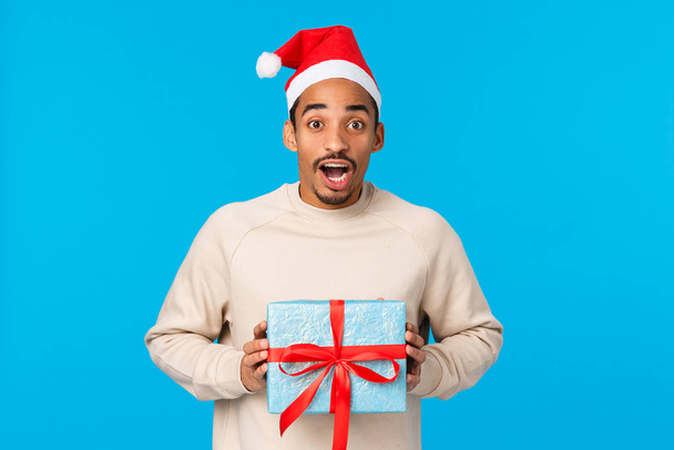 Здивований захоплений і переповнений афро-американський милий чоловік у капелюсі Санта, різдвяний наряд, тримає подарунок на Новий рік від друга і відчуває себе розважальним, хоче неперевершений подарунок, синій фон
 - Фото, зображення