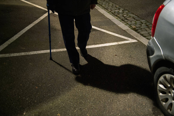 Σιλουέτα και σκιά του ηλικιωμένου άνδρα που περπατούσε στο γαλλικό δρόμο τη νύχτα χρησιμοποιώντας μπαστούνι που διατηρεί την ισορροπία με μπλε τηλεσκοπικό μπαστούνι αλουμινίου Ιατρική βοήθεια και αποκατάσταση - Φωτογραφία, εικόνα