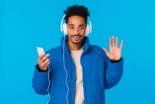 Hey nice see you. Cheerful happy stylish african-american man in padded winter jacket, listen music headphones, hold smartphone switch song, waving camera hello, hi gesture, blue background - Fotografie, Obrázek