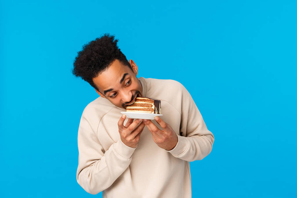 Guy stole piece cake and eating it fast. Funny and cute african-american man holding plate, bite delicious dessert celebrating birthday, enjoying holidays tasty food, standing blue background - Foto, imagen