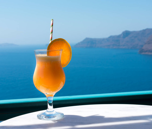 Friss narancslevet facsart, készen a részegségre. A narancslevet egy átlátszó üvegpohárban, szívószállal tálalják fel az asztalon, és narancs szelettel díszítik. Az Égei-tenger a háttérben - Fotó, kép