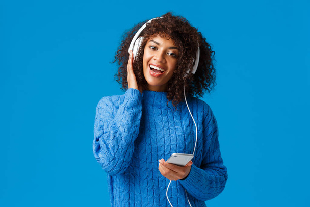 Heureuse et joyeuse femme afro-américaine jouant à l'application de jeu de karaoké sur smartphone, portant des écouteurs chantant et appréciant une musique impressionnante, une qualité sonore, un fond bleu debout
 - Photo, image