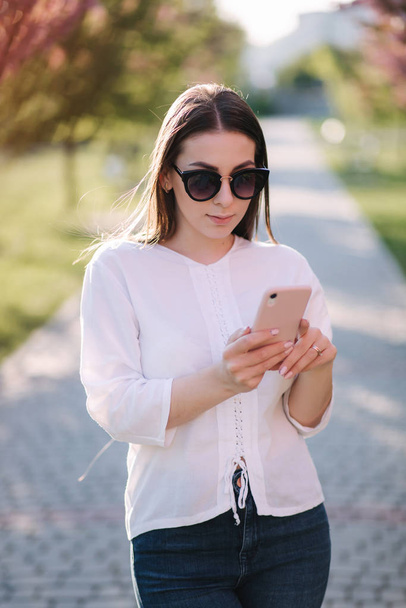 Portrét attravní mladé ženy v bílé košili a slunečních brýlích používejte venku smartphone. SPEEL s někým v telefonu. Pozadí růžového stromu - Fotografie, Obrázek