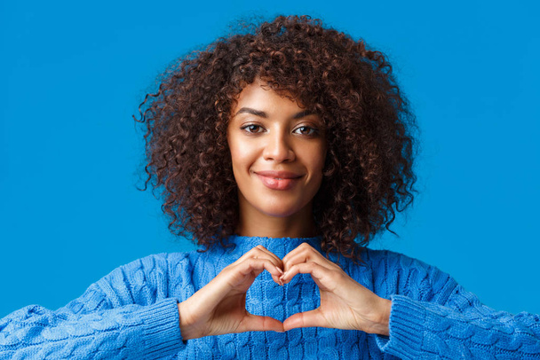 Портрет крупным планом нежная и милая афро-американская романтическая женщина выражает свои чувства жестом, показывая знак сердца и улыбаясь, сочувствуя, признаваясь на синем фоне
 - Фото, изображение