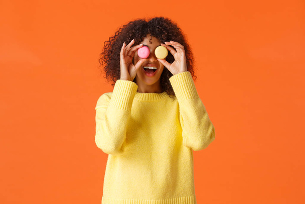 Hravé legrační a bezstarostné, hezká africko-americká kudrnatá dívka ve žlutém svetru, blázen kolem dělat oči z makaronů a usmívat se, jíst sladkosti, jako dezerty, stojící oranžové pozadí - Fotografie, Obrázek