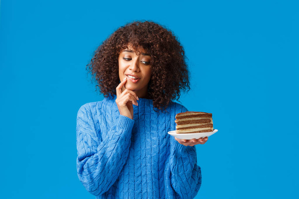 Chica quiere comer delicioso pedazo de pastel, pero preocuparse por la dieta y las calorías. Atractiva tonta mujer afroamericana tratando de resistir la tentación tomar bocado de postre sabroso, mirando con deseo en el plato
 - Foto, Imagen