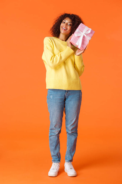Full-length κάθετη βολή χαρούμενη χαρισματική αφροαμερικάνικη γυναίκα κουνώντας κουτί με το παρόν να μαντέψει τι είναι μέσα, χαμογελώντας κρατώντας τυλιγμένο δώρο κοντά στο πρόσωπο, στέκεται πορτοκαλί φόντο - Φωτογραφία, εικόνα