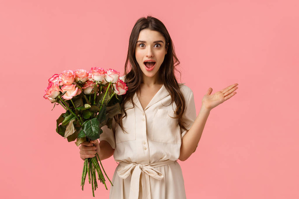 Excited and amused, surprised gorgeous modern woman in trendy dress, spread hands sideways amazed and astonished, look astounded by nice gesture, holding pretty flowers, roses bouquet - Foto, Imagen