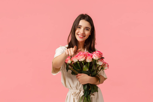 Ты заслуживаешь лучшего. Очаровательная милая и романтичная, нежная кавказская девушка в платье, держа розы возле груди, аккуратно оберните букет и укажите пальцем камеру, улыбаясь радостно, стоя розовый фон
 - Фото, изображение