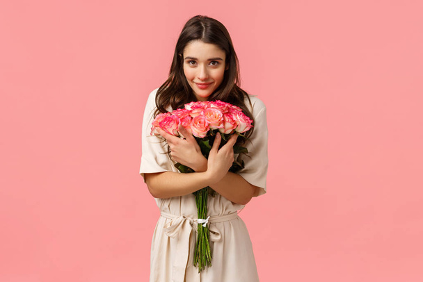 Tenderness, romance and love concept. Attractive brunette female, girlfriend receiving beautiful flowers, hugging bouquet and smiling camera delighted, show affection and happiness, pink background - Photo, Image