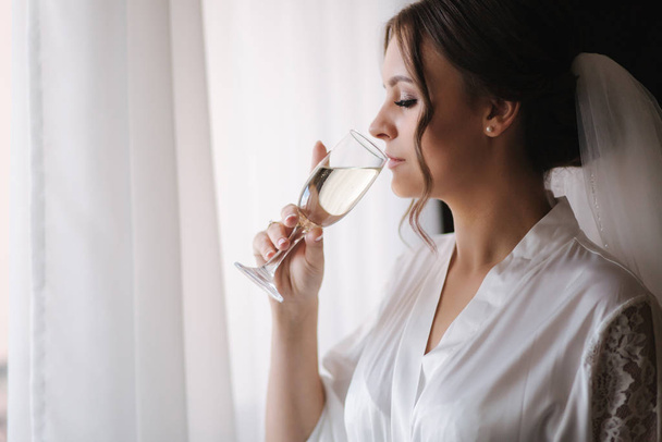 Portrait of gorgeous bride at hotel in morning. Charming woman with glass of champagne - Foto, Bild
