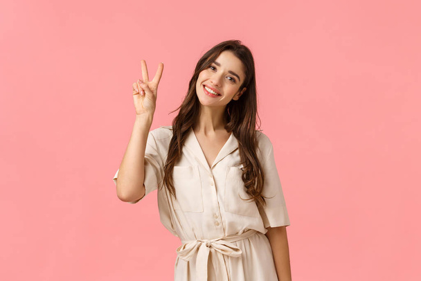 Women, beauty and advertising concept. Cheerful tender brunette woman feeling relaxed and delighted, smiling lovely, tilt head coquettish as wearing dress showing peace sign, pink background - Foto, Imagem