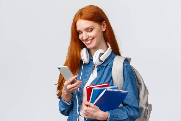 Teenagers, college and education concept. Attractive redhead female, univeristy student with headphones, backpack and notebooks, answer girlfriend message, queer love in air - Photo, Image