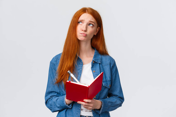 Uneasy and gloomy, indecisive cute redhead girl writing down her feelings in red notebook, looking nervous and troubled up thinking, holding pen, pondering something while sharing thoughts with paper - Photo, Image