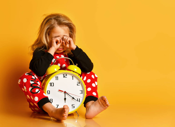 Little blond girl in red dotted pajamas sitting on floor with big alarm clock, feeling tired, yawning and rubbing eyes - Photo, image