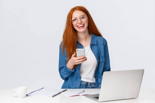 Cheerful, friendly-looking charismatic redhead female designer, freelancer with red foxy hair, glasses, laughing carefree, holding smartphone, working over project with laptop, drink coffee - Photo, Image