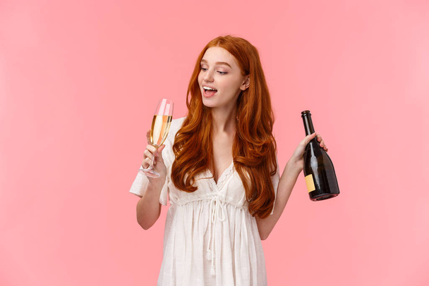 Party, hungover and celebration concept. Happy and cheerful, carefree good-looking redhead woman drinking champagne from glass, holding bottle, got drunk, wasted standing over pink background - Photo, Image