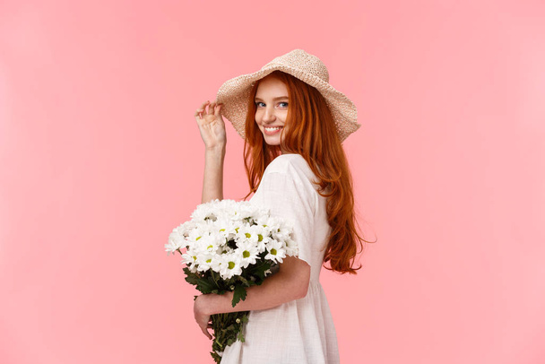 Romantic, silly and tender feminine redhead woman in cute hat, dress, holding bouquet white flowers, turn camera and smiling coquettish, flirting with boyfriend over pink background - Foto, afbeelding