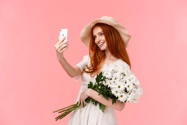 Celebración, redes sociales y concepto de internet. Atractivo atrevida pelirroja hembra en sombrero de paja, vestido de primavera, la celebración de ramo, tomar selfie en el teléfono inteligente con flores blancas, sonriendo complacido
 - Foto, Imagen