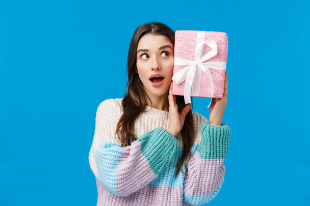 Waist-up portrait curious attractive young woman 20s, brunette shaking cute present to guess what inside, feeling intrigued receiving christmas gift, standing wondered and amused blue background - Photo, Image