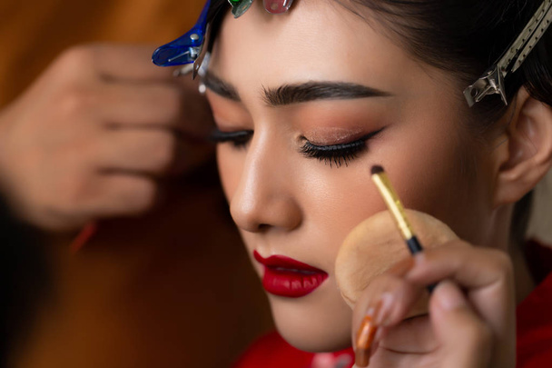 Mooie Aziatische jonge bruid toe te passen mooie make-up en kapsel door make-up kunstenaar, hairstylist voor huwelijksceremonie. Aantrekkelijke aziatische vrouw krijgen mooie rode lippenstift, wimper verlengt, dikke wenkbrauwen - Foto, afbeelding