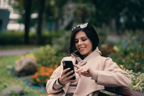 ベンチに座って街の公園でスマートフォンを使って幸せな女の子 - 写真・画像