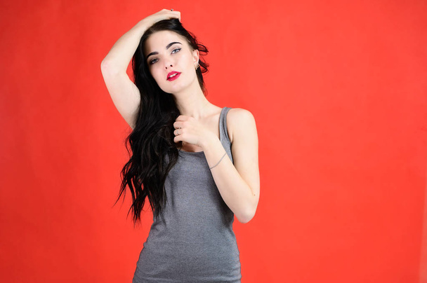 The concept of a beautiful female portrait with emotions. Portrait of a brunette girl with a smile with long hair with excellent makeup in a gray dress on a red background in different poses. - Foto, Bild