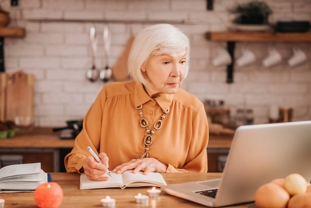Grey-haired beautiful elderly lady in orange blouse reading a lesson online - Фото, зображення
