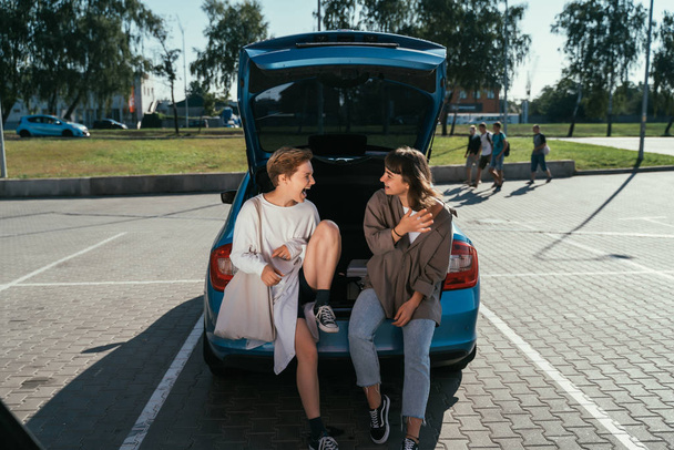 Zwei Mädchen posieren auf dem Parkplatz am offenen Kofferraum für die Kamera. - Foto, Bild