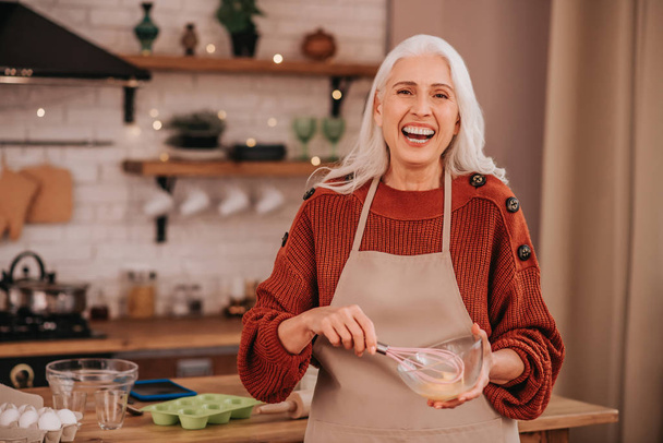 Grey-haired smiling lady smiling bright whipping milk in a bowl - Fotografie, Obrázek