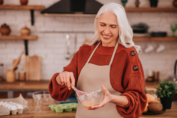 Grey-haired smiling lady whipping milk in a bowl - Photo, Image