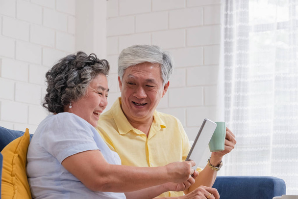 Ασιατικό ζευγάρι ηλικιωμένων χρησιμοποιώντας πίνακα και να πίνουν καφέ που κάθονται στον καναπέ στο σαλόνι στο σπίτι. ανώτερη με την τεχνολογία της τεχνολογίας. γήρανση στο σπίτι έννοια. - Φωτογραφία, εικόνα