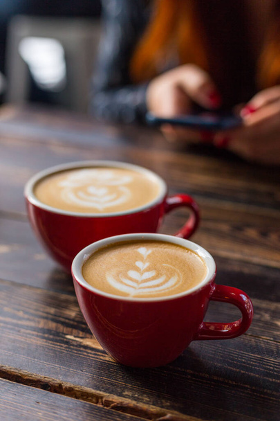 Δύο κόκκινα φλιτζάνια καφέ σε ξύλινο φόντο με latte art. Τραπέζι στο καφέ. Έννοια του εύκολου πρωινού. Μικρά και μεγάλα κεραμικά κύπελλα. Στο πάνω μέρος. Κορίτσι με ένα τηλέφωνο στο παρασκήνιο - Φωτογραφία, εικόνα