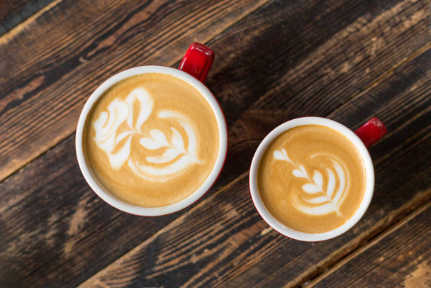 Two red cups of coffee on wooden background with latte art. Table in cafe. Concept of easy breakfast. Small and big ceramic cups. Top view - Photo, Image