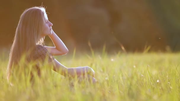 Νεαρή γυναίκα με μακριά μαλλιά κάθεται σε εξωτερικούς χώρους το καλοκαίρι γρασίδι απολαμβάνοντας τη φύση στο ηλιοβασίλεμα. - Πλάνα, βίντεο