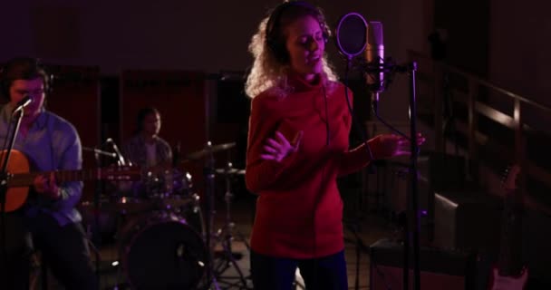  Caucasian female singer with blonde curly hair wearing headphones and singing into a microphone, a Caucasian male singer guitarist and a male drummer in the background, performing during a session at a recording studio - Video, Çekim