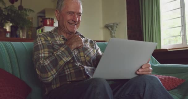 Front view of a senior Caucasian man relaxing at home in his living room, sitting on the sofa using a laptop computer and laughing, slow motion - Metraje, vídeo