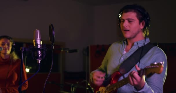 Caucasian man wearing headphones singing into a microphone and playing electric guitar with a Caucasian woman singing and a drummer playing in the background, in a live recording room at a recording studio. Musicians working on producing a song - Кадры, видео