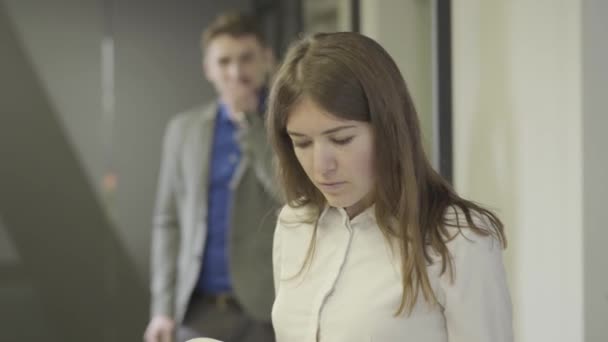 Close-up of beautiful young Caucasian woman looking through documents as handsome man checking his suit at the background and coming up. Office romance, flirt, flirting. - Imágenes, Vídeo