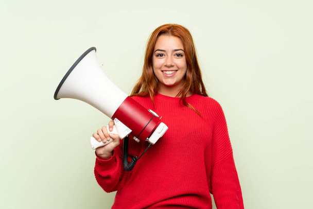 Adolescente rousse fille avec pull sur fond vert isolé tenant un mégaphone
 - Photo, image