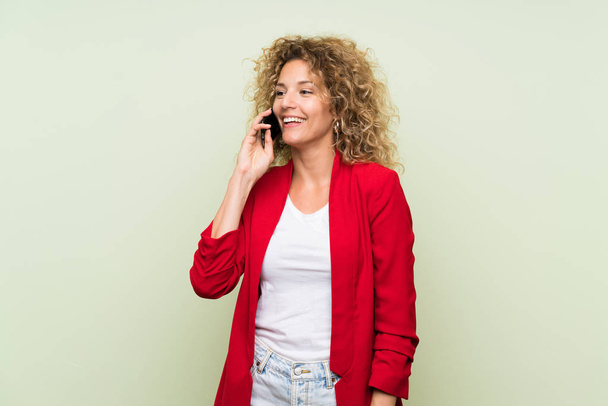 Young blonde woman with curly hair over isolated green background keeping a conversation with the mobile phone - 写真・画像