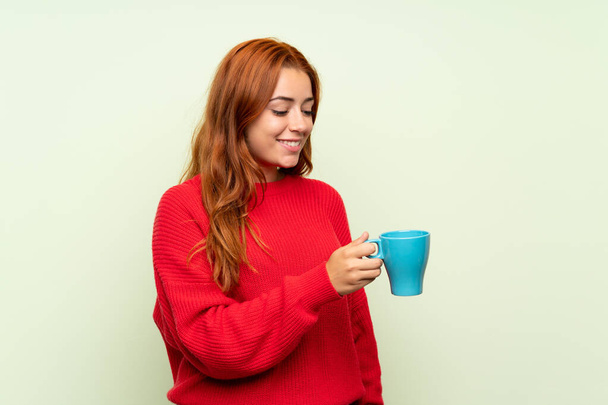 Tinédzser vöröshajú lány pulóvert több mint elszigetelt zöld háttér gazdaság forró csésze kávét - Fotó, kép