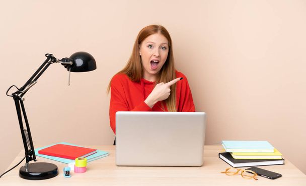 Young student woman in a workplace with a laptop surprised and pointing side - Photo, Image